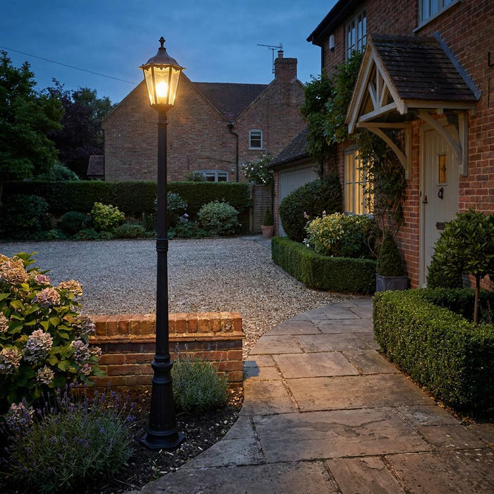 Traditional black lamppost lighting a stone path and gravel driveway in front of a brick house at dusk.