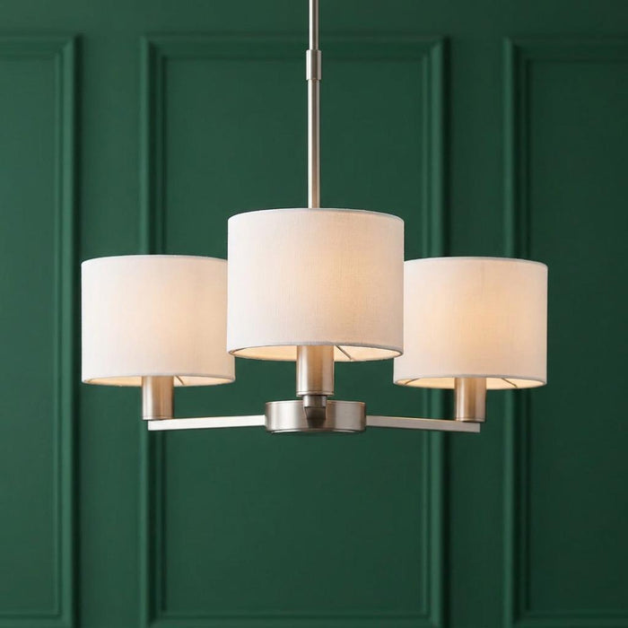 Three-light satin nickel ceiling pendant with white drum shades against a dark green paneled wall.