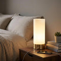 Cylindrical brass table lamp with a glowing white glass shade on a wooden bedside table.