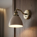 Antique brass wall light with a taupe dome shade, mounted on dark wood and glowing with warm light.