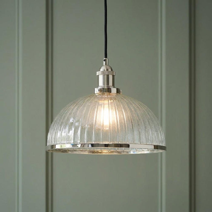 Lit industrial pendant light with a ribbed glass shade and polished nickel trim against a sage green paneled wall.