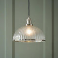 Lit industrial pendant light with a ribbed glass shade and polished nickel trim against a sage green paneled wall.