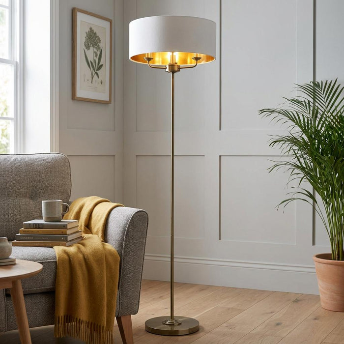 Antique brass floor lamp with a white drum shade and gold interior, standing beside a grey armchair.