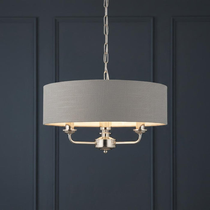 A bright nickel three-light pendant with a charcoal fabric drum shade against a dark blue paneled wall.