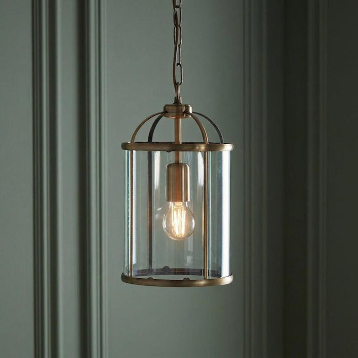 Antique brass lantern pendant light with clear glass and a glowing bulb, hanging against a paneled wall.