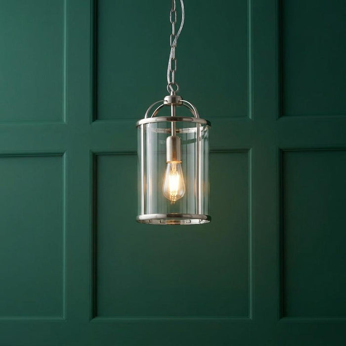Polished nickel ceiling lantern with clear glass shade and warm bulb glowing against a dark green paneled wall.
