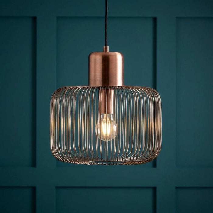 Antique copper wire cage pendant light with a glowing bulb against a teal paneled wall.