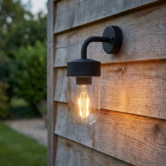 Modern black outdoor wall light with a glass capsule shade and a lit filament bulb on a wooden exterior wall.