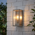 Rectangular brushed stainless steel outdoor wall light with a glowing filament bulb mounted on a white brick wall.