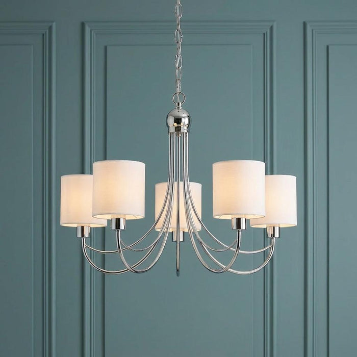 Chrome five-arm chandelier with white cylinder shades against a teal paneled wall.
