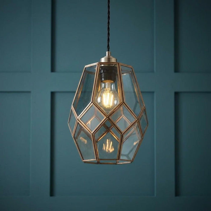 Geometric brass and glass pendant shade illuminated by an Edison bulb against a dark teal paneled wall.