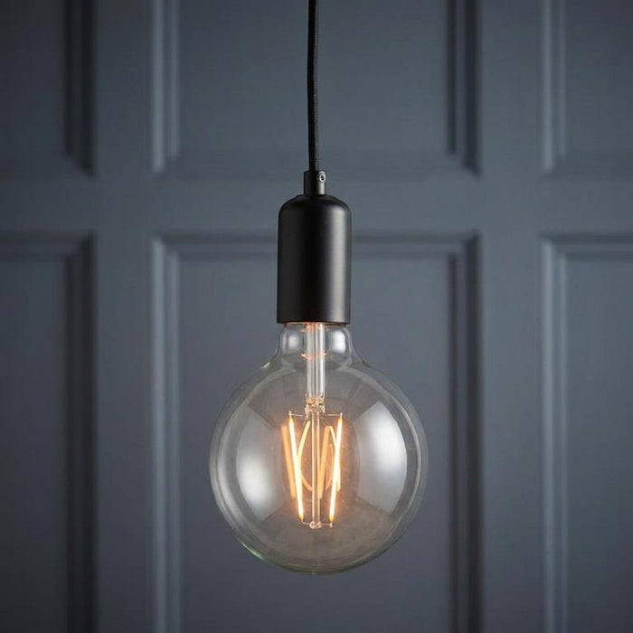 Minimalist black pendant light with a large clear spherical filament bulb against a dark blue paneled wall.
