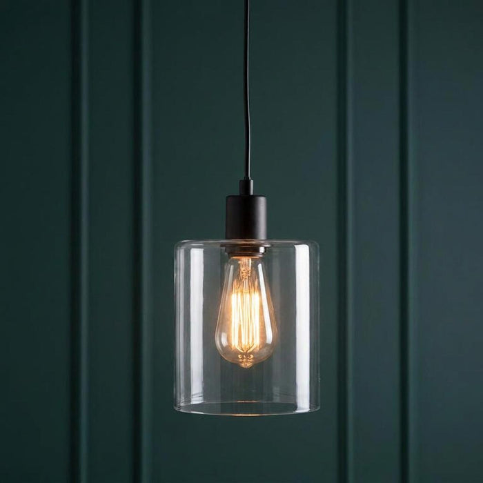 Black pendant light with a clear cylindrical glass shade and Edison bulb glowing against dark green paneling.