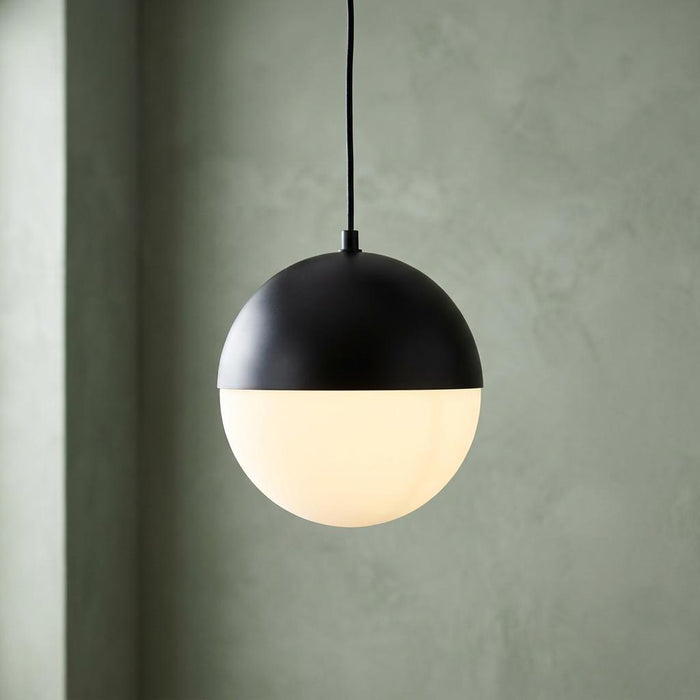 Spherical pendant light with a matte black top and glowing opal glass base against a textured sage green wall.