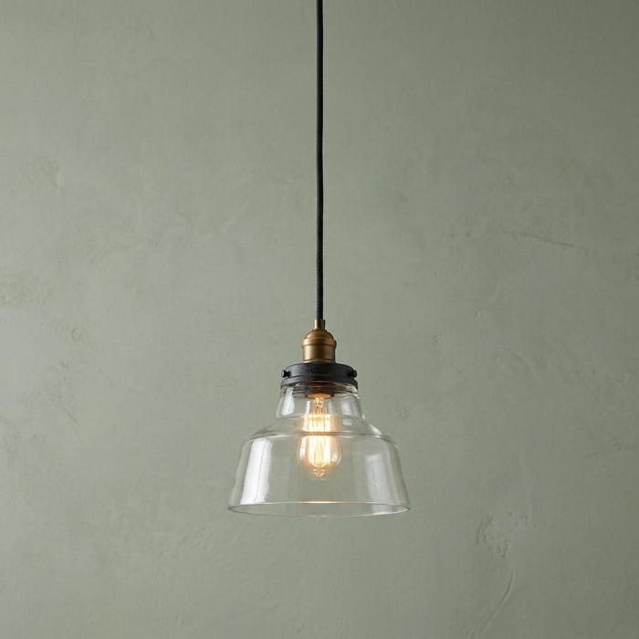 A Feiss Baskin 1 light aged brass and zinc ceiling pendant with a clear glass shade against a sage green wall.