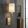 Feiss Brianna brass wall sconce with a white linen cylindrical shade mounted on a textured grey wall.
