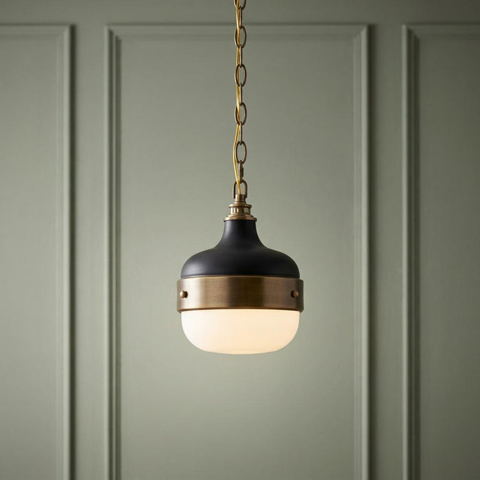 Feiss Cadence mini pendant light with black and brass metalwork and opal glass, hanging against a green paneled wall.