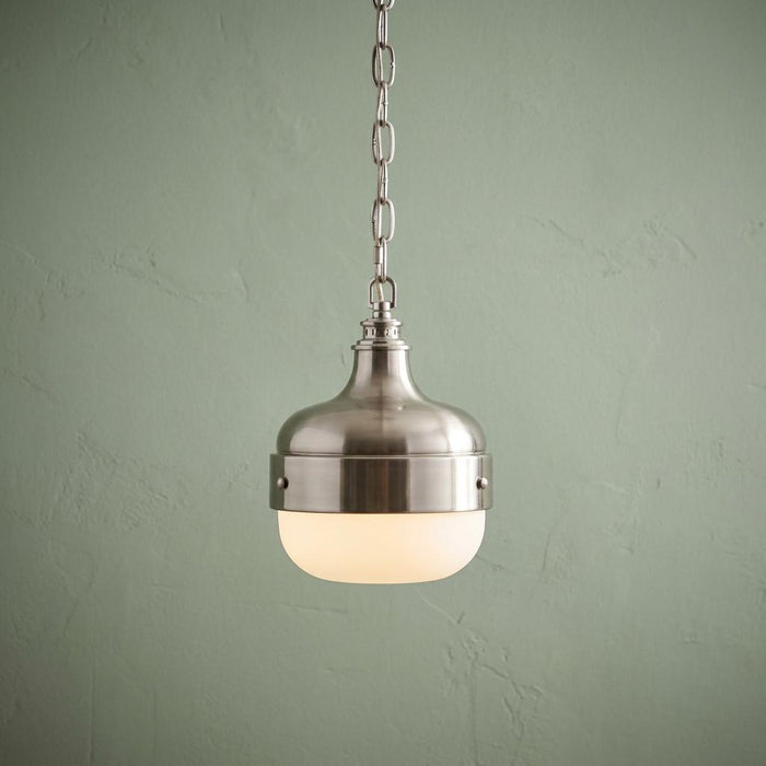 Brushed steel mini pendant light with a warm glowing opal glass shade against a pale green textured wall.