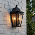 Black traditional outdoor half wall lantern with two lit bulbs mounted on a white brick wall.