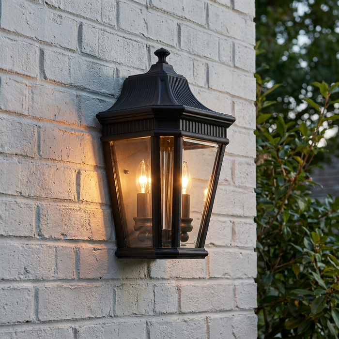 Black traditional outdoor half wall lantern with two lit bulbs mounted on a white brick wall.