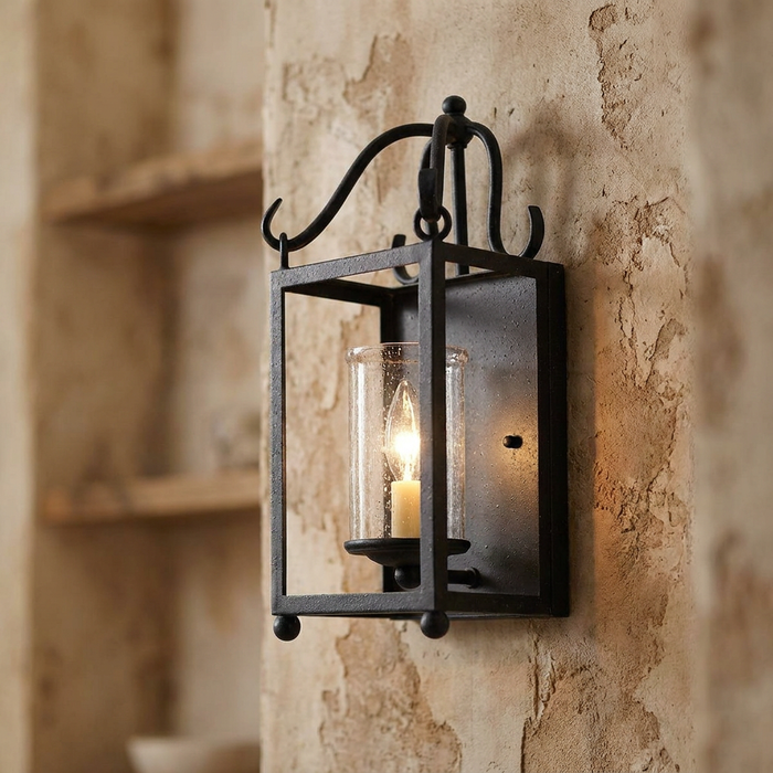 Antique iron wall lantern with a lit candle bulb in a glass cylinder, mounted on a rustic stone wall.