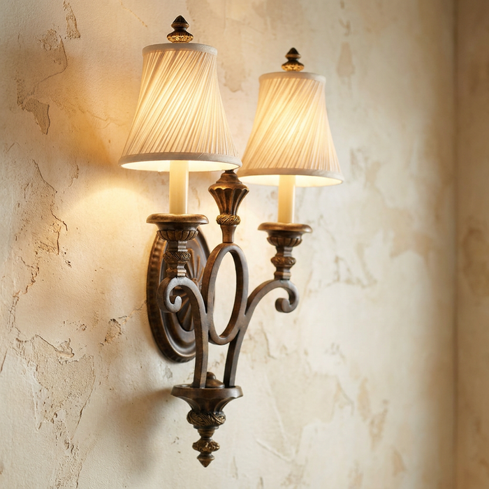 Twin arm wall sconce with walnut finish and pleated shades emitting a warm glow on a textured plaster wall.