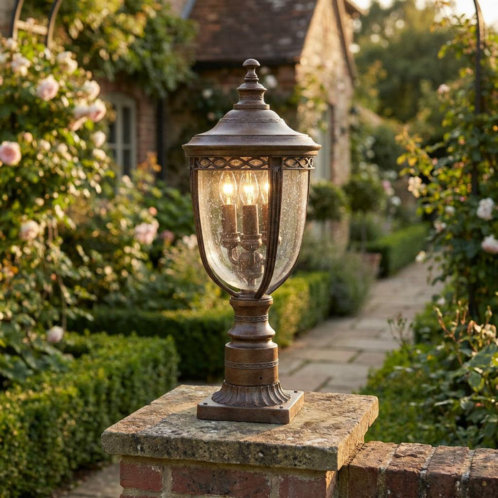 Weathered bronze pedestal lantern with seeded glass glowing on a stone garden pillar amidst blooming roses.