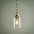 Brass mini pendant light with a clear glass cylinder shade and a lit bulb hanging against a pale green wall.