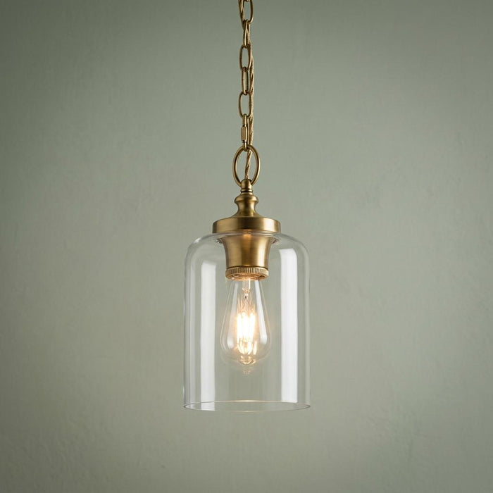 Brass mini pendant light with a clear glass cylinder shade and a lit bulb hanging against a pale green wall.
