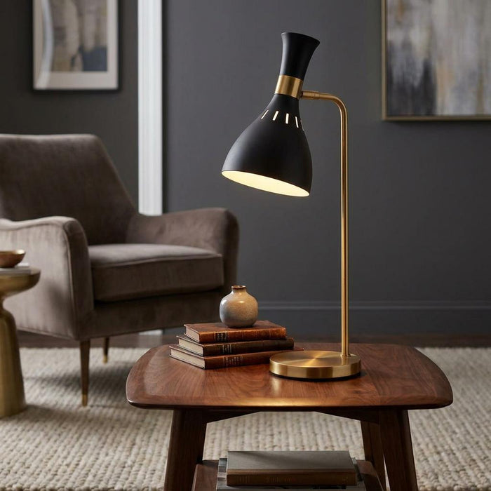 Black and brass table lamp with an hourglass shade illuminated on a wooden side table in a cozy living room.