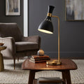 Black and brass table lamp with an hourglass shade illuminated on a wooden side table in a cozy living room.