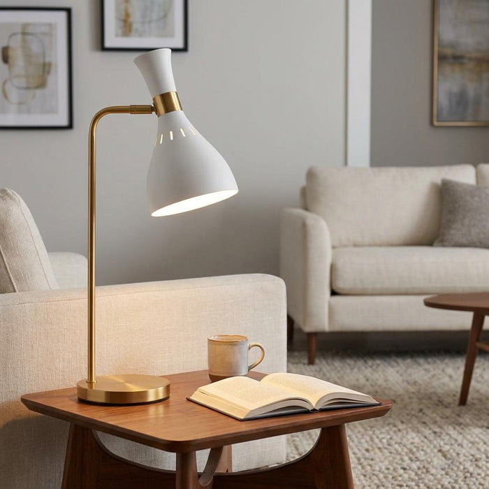 Matte white and brass table lamp casting light on an open book in a contemporary living room.