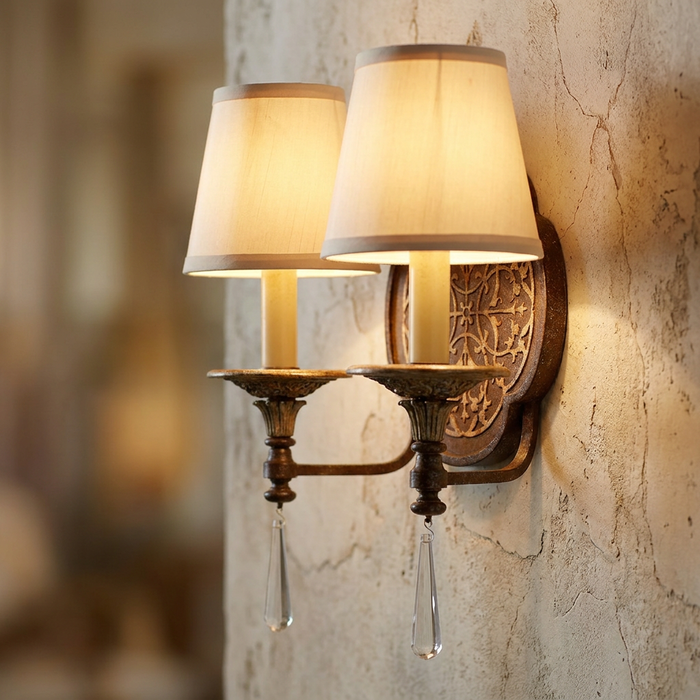 Two-light bronze wall sconce with cream shades and crystal droplets on a rustic textured wall.