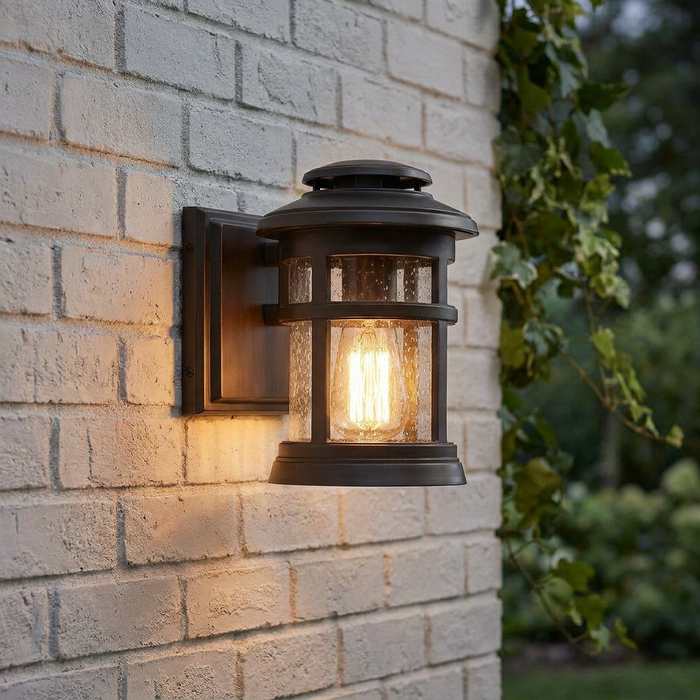 Bronze outdoor wall lantern with seeded glass and a glowing Edison bulb mounted on a light brick wall.