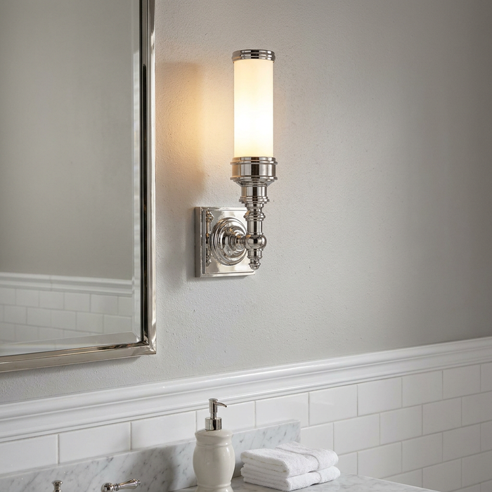 Ornate chrome wall sconce with a cylindrical white shade mounted in a bathroom next to a mirror.