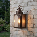 Copper outdoor wall lantern with seeded glass panels and a lit bulb mounted on a white brick wall.
