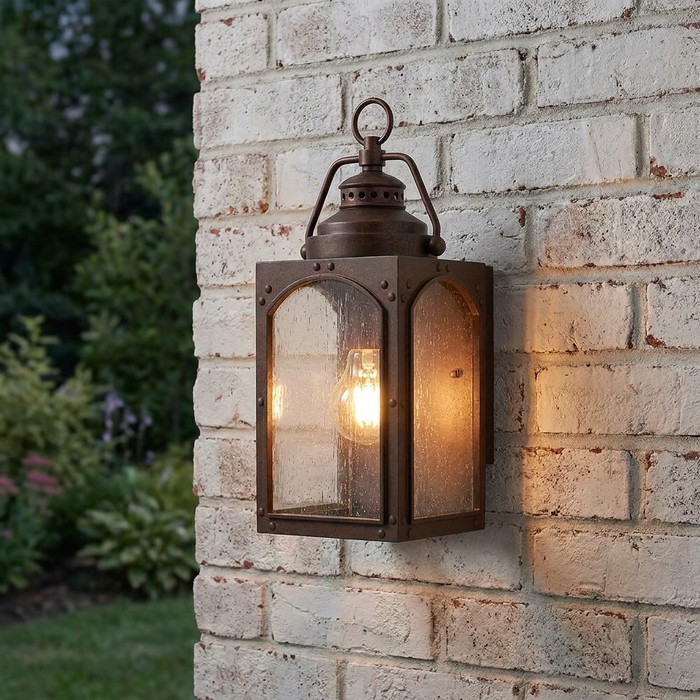 Copper outdoor wall lantern with seeded glass panels and a lit bulb mounted on a white brick wall.