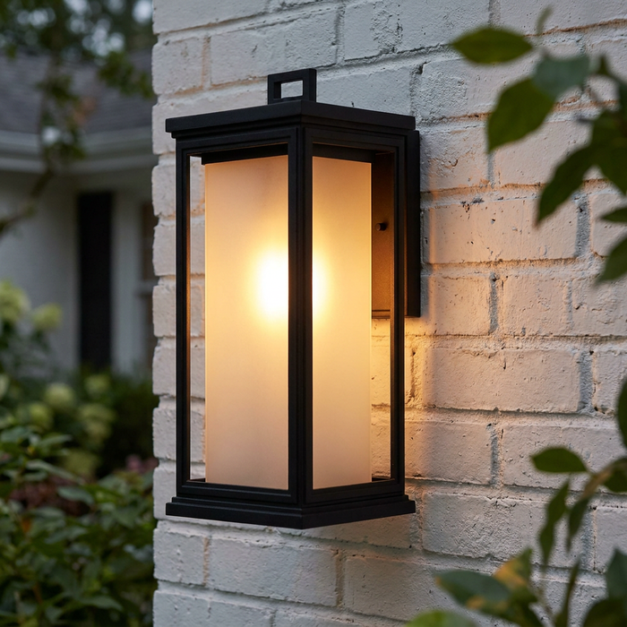 Black lantern wall light with a frosted glass interior glowing on a white brick wall.