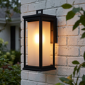 Black lantern wall light with a frosted glass interior glowing on a white brick wall.