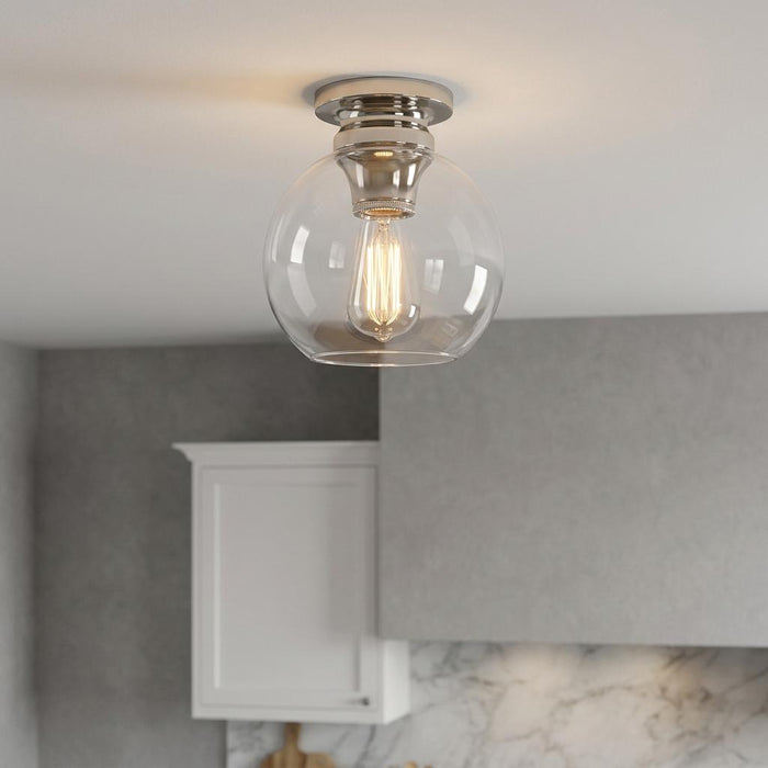 Polished nickel flush mount ceiling light with a clear glass globe and glowing Edison bulb in a modern interior.