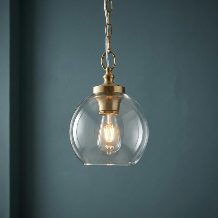 Feiss Tabby mini pendant light with a clear glass globe shade and brass hardware, glowing against a dark teal wall.