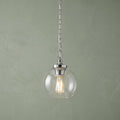 A Feiss Tabby polished nickel pendant light with a clear glass globe and glowing bulb against a green wall.