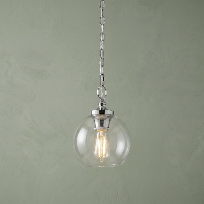 A Feiss Tabby polished nickel pendant light with a clear glass globe and glowing bulb against a green wall.