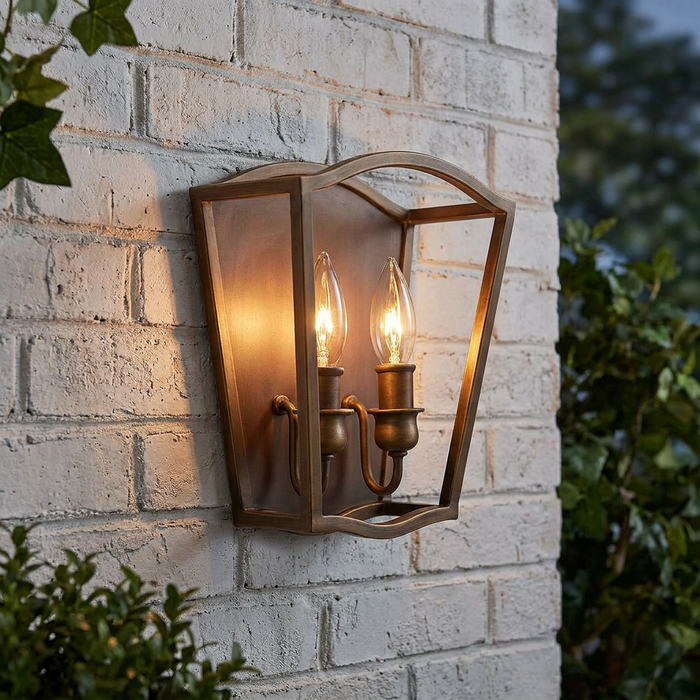 Two-light aged brass wall lantern with glowing candle bulbs mounted on a white brick wall.