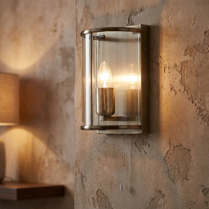 Fern antique brass wall light with curved glass and a lit bulb mounted on a rustic textured wall.