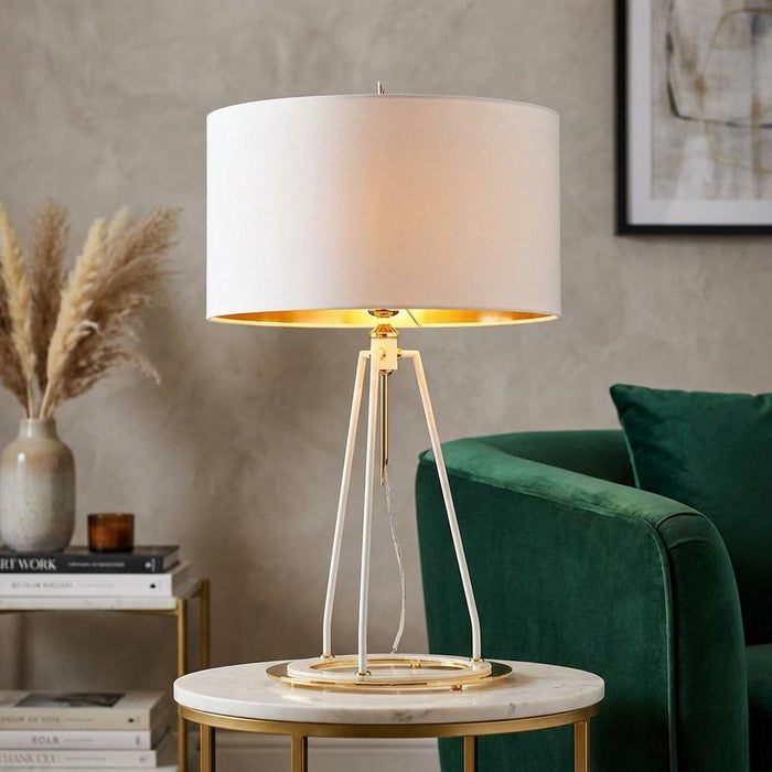 White drum-shaded lamp with a polished gold tripod base on a marble table next to a green velvet armchair.