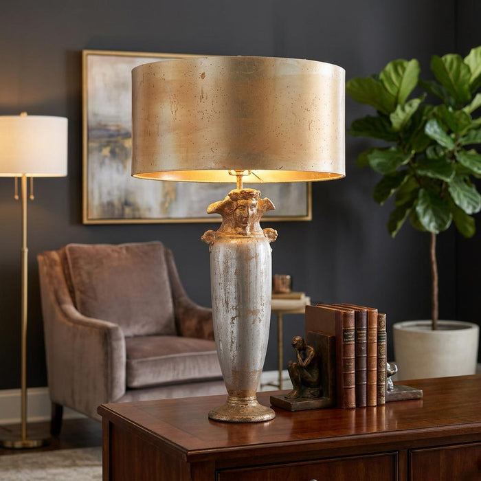 Ornate silver and gold table lamp with a sculpted urn base and metallic drum shade in a refined interior setting.