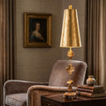 Gold leaf table lamp with a sculptural base of classical fragments sits beside an armchair in a warm room.