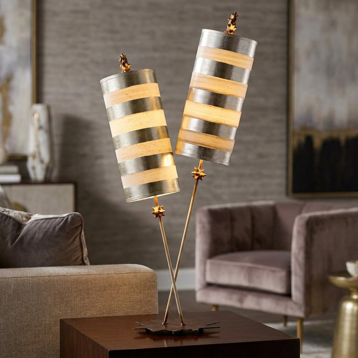 Modern table lamp with two striped silver and cream cylindrical shades on thin crossing stems in a living room.