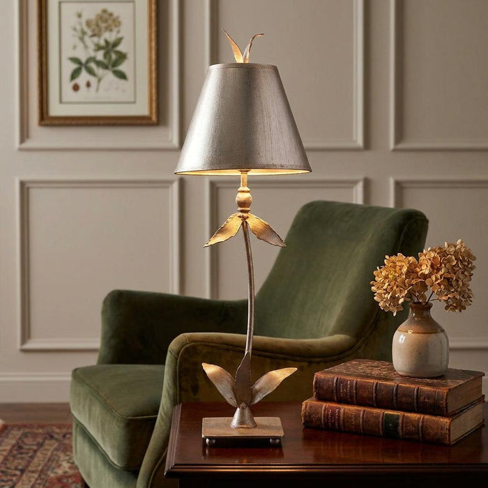 Silver leaf table lamp with a floral stem and metallic shade beside a green velvet armchair.